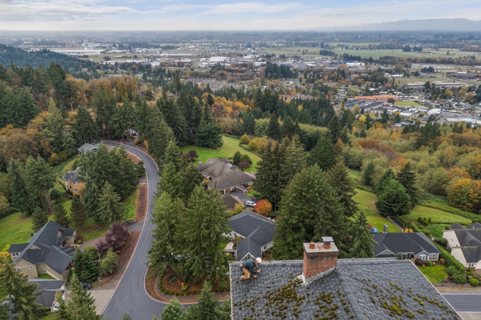 Roofing contractor inspecting deteriorated step flashing and moss-covered asphalt shingles on a ranch-style home under an overcast Pacific Northwest sky
